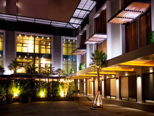 Night exterior of a multi-storey hotel building with illuminated windows, palm trees, uplit foliage and a covered walkway with a corrugated roof.
