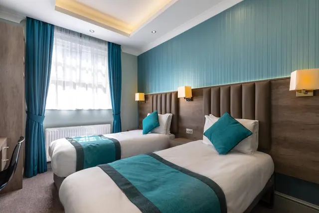 A twin bedroom with two single beds with teal throws and cushions, a window with blue curtains, and wall lights above the headboards.