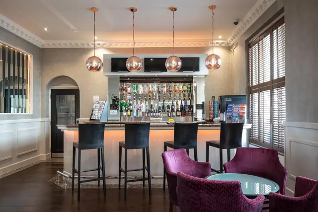 A hotel bar with a backlit counter, dark bar stools, copper pendant lights and purple armchairs around a round table.