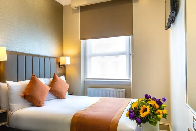 A double bed with white linen and orange cushions sits beside a window with a brown roller blind, with a wall-mounted television and a vase of flowers on the right.