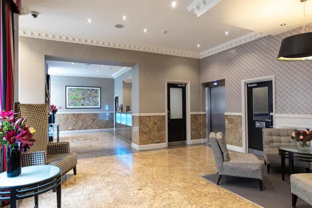 A hotel lobby with upholstered armchairs, a round side table holding flowers, a lift door and a reception desk in the background.