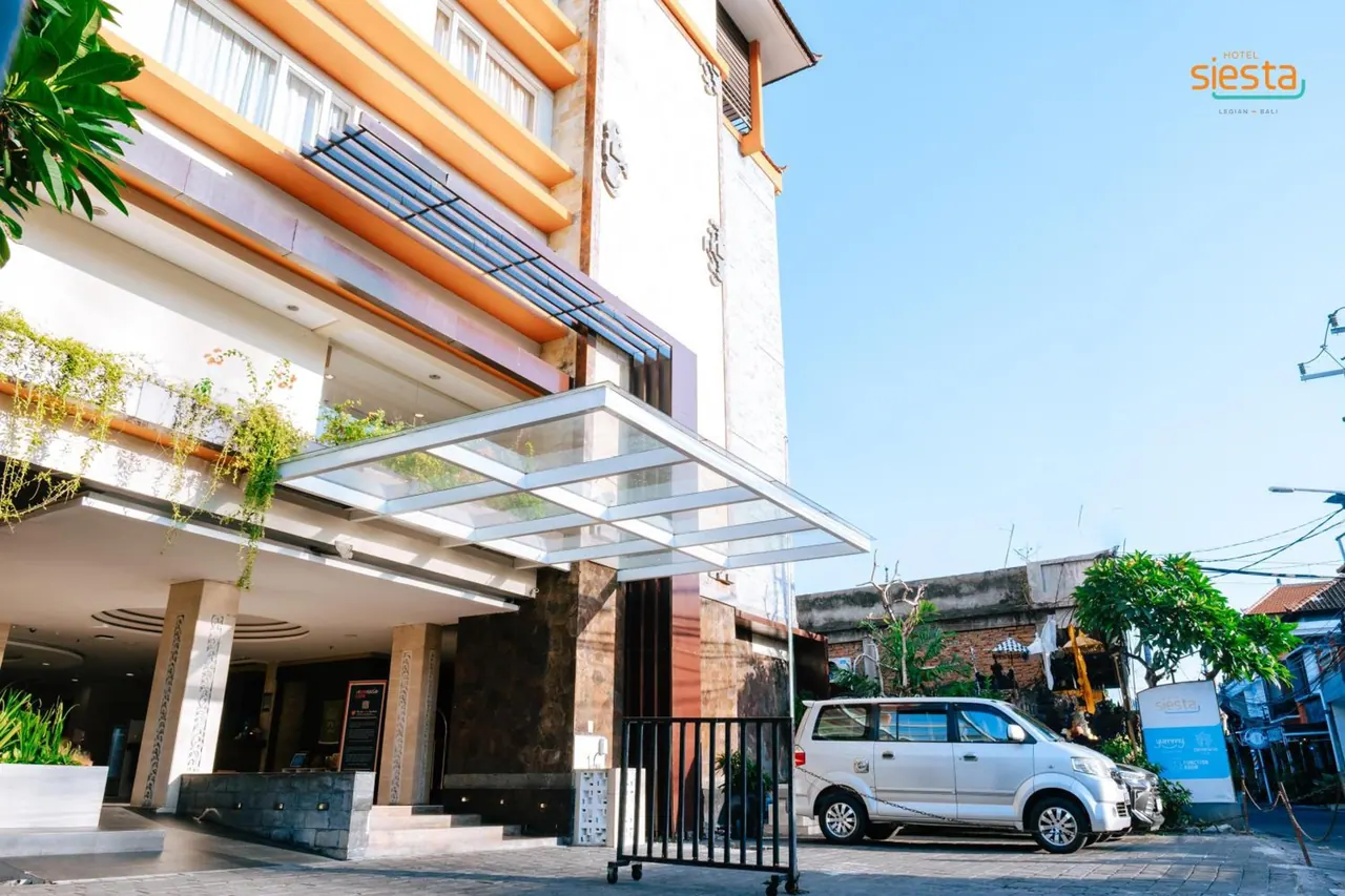 The front of Hotel Siesta shows a glass canopy over the entrance, a white minivan parked at the side and a clear blue sky above.