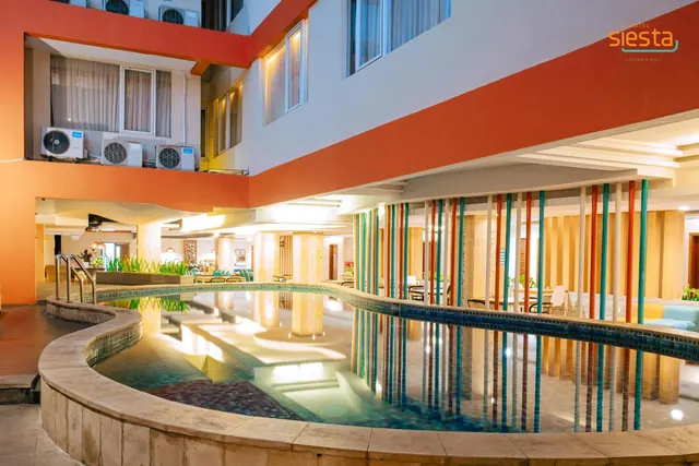 An indoor pool with a curved tiled edge and vertical colourful poles in an atrium, with air conditioning units on the wall above and a hotel logo in the top right corner.