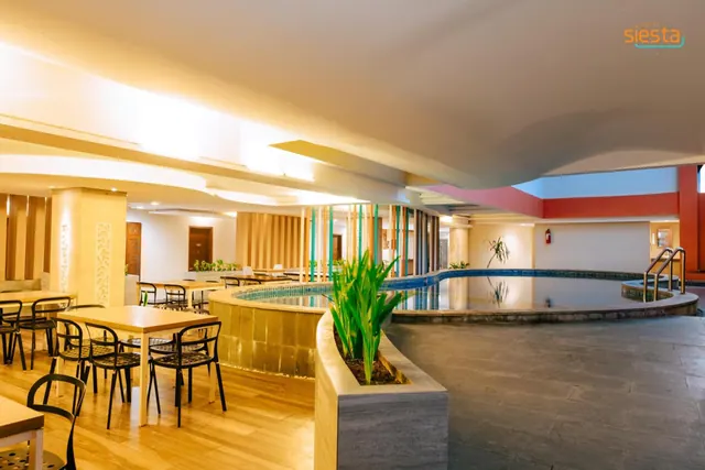 An indoor pool with a tiled edge and metal handrails sits beside a dining area with wooden tables and chairs under warm ceiling lights.