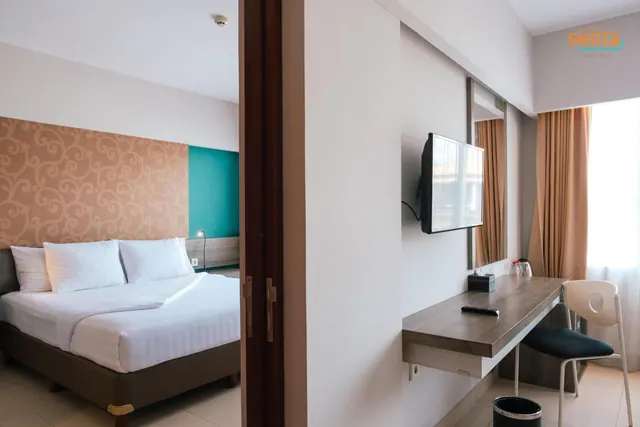 A bedroom with a made bed with white linen, a wall-mounted television, a wooden desk with a chair, and a window with curtains, with a hotel logo watermark in the corner.