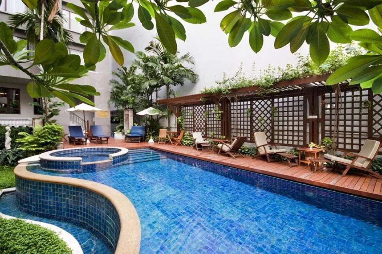 An outdoor pool with blue mosaic tiles, a small round spa pool, a wooden deck with sun loungers and white parasols, and tropical plants around it.