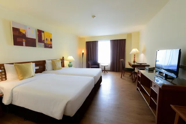 A hotel bedroom with two single beds with white bedding, a wooden desk and chair, an armchair by the window, and a wall-mounted flat-screen television on a wooden cabinet.