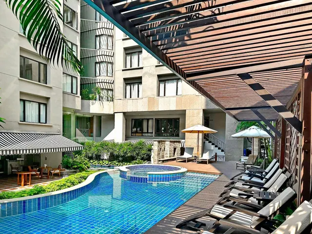 A blue-tiled swimming pool with a small round jacuzzi beside it, surrounded by sun loungers under a wooden pergola, with hotel building walls behind.