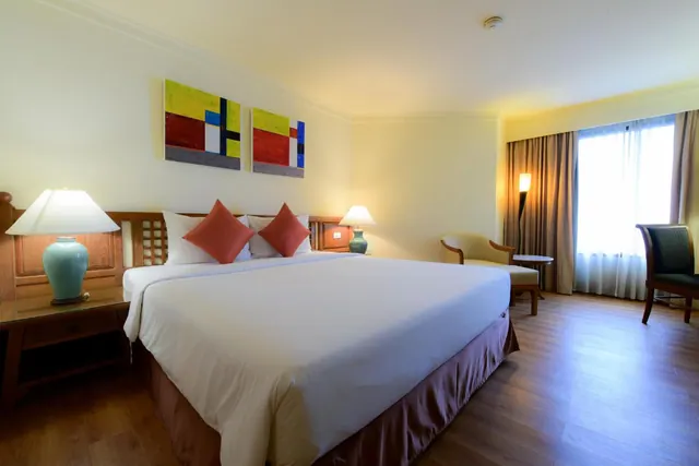 A bedroom with a large white bed, red cushions, two teal table lamps, abstract wall paintings, and an armchair beside a window with sheer curtains.