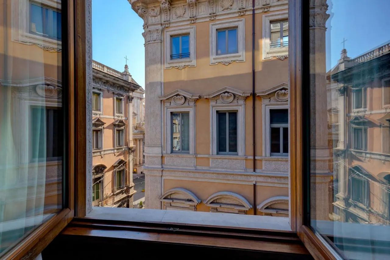 View through an open wooden-framed window across a narrow street to ochre palazzo facades with ornate window surrounds under a blue sky.