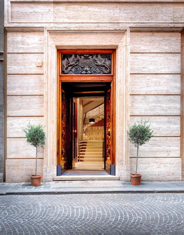 A travertine facade with an open wooden doorway under a carved panel reading SINGER, a staircase visible inside, and two potted olive trees on the pavement outside.