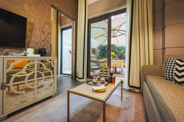 A living area with a sofa, coffee table, wall-mounted television and a sideboard holding a kettle, with glass doors opening onto a terrace with an umbrella, table and greenery.
