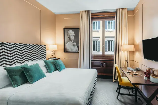 A bedroom with a made double bed with teal cushions, a framed black and white bust photograph, a window onto a building facade, and a desk with a yellow chair and a wall-mounted television.
