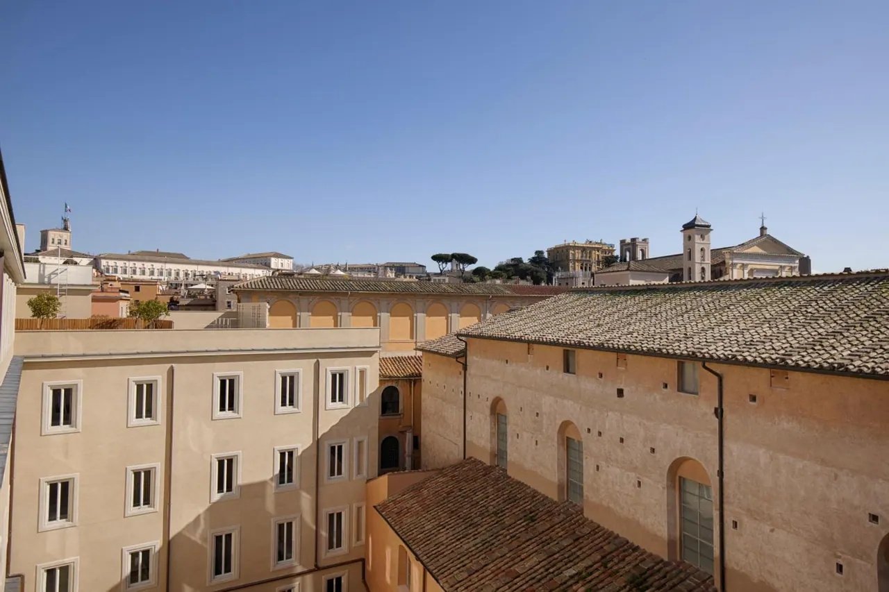 Looking out over terracotta tiled rooftops and ochre building facades toward a church with a bell tower and pine trees under a clear blue sky.
