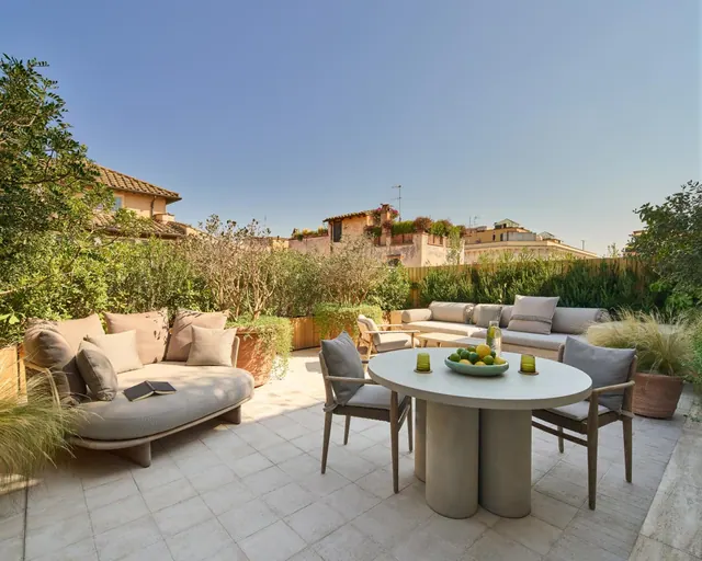 A furnished outdoor terrace with sofas, a round dining table and chairs, and potted plants, with rooftops and buildings visible beyond the hedge.