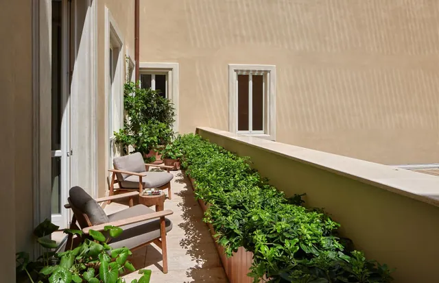 A narrow sunlit balcony with two wooden armchairs with grey cushions, a small side table, and planters of green shrubs along the railing, facing a beige building wall.