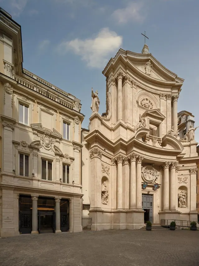 Exterior view of a baroque church facade with columns and statues beside a stone palazzo with a hotel entrance on a cobbled square.