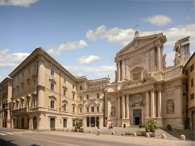 The photo shows the exterior of a cream-coloured palazzo beside a baroque church facade on a sunny street, with a few people walking across the square.