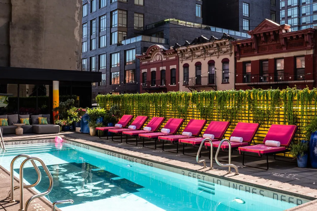 A rooftop swimming pool with pink sun loungers beside a green climbing hedge, with city apartment buildings and historic brick townhouses behind.