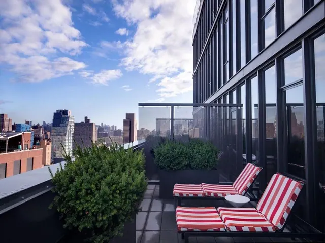 A rooftop terrace with red and white striped loungers, potted shrubs and a glass railing, overlooking city buildings under a partly cloudy sky.