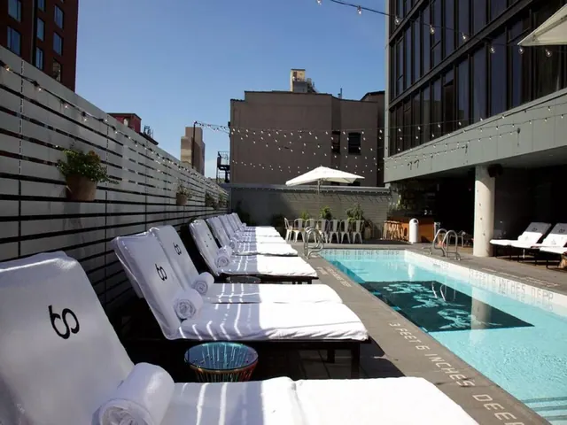 A rooftop pool with white lounge chairs and rolled towels along one side, a white umbrella and string lights, and neighbouring buildings behind.