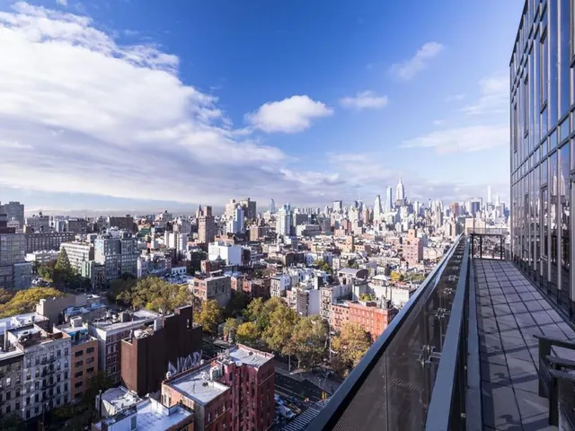 A rooftop terrace with a glass railing overlooks a dense city skyline under a partly cloudy blue sky, with the Empire State Building visible in the distance.