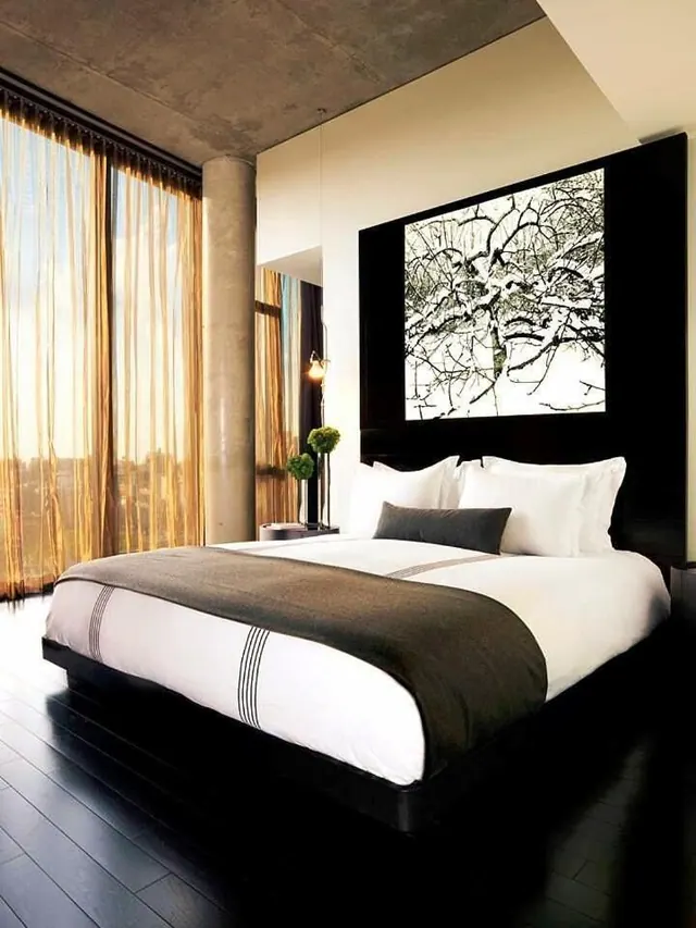A king bed with white linens and a dark headboard sits in a bedroom with floor-to-ceiling sheer curtains and a hazy city view through the window.