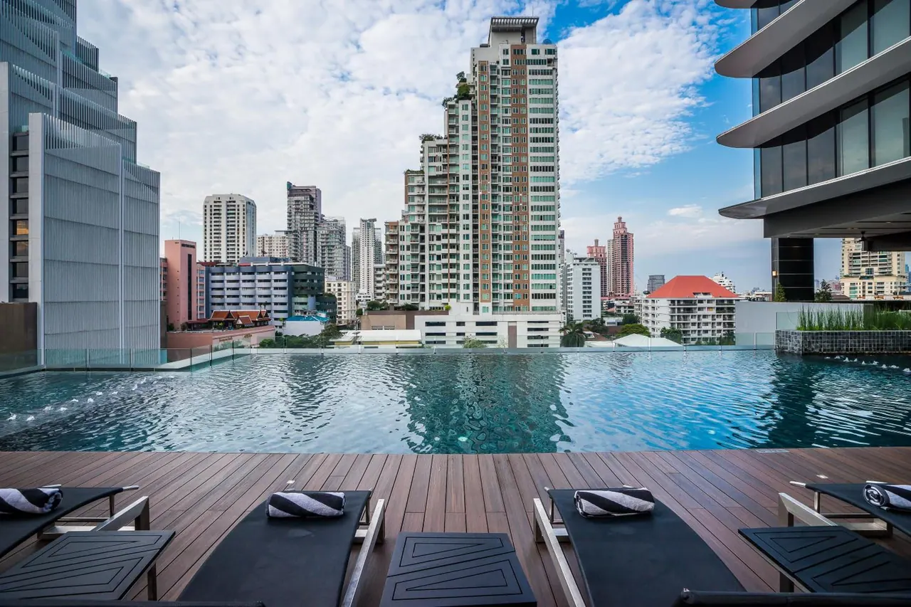 An infinity edge rooftop pool with sun loungers and folded towels on a timber deck, overlooking a city skyline of high-rise buildings.