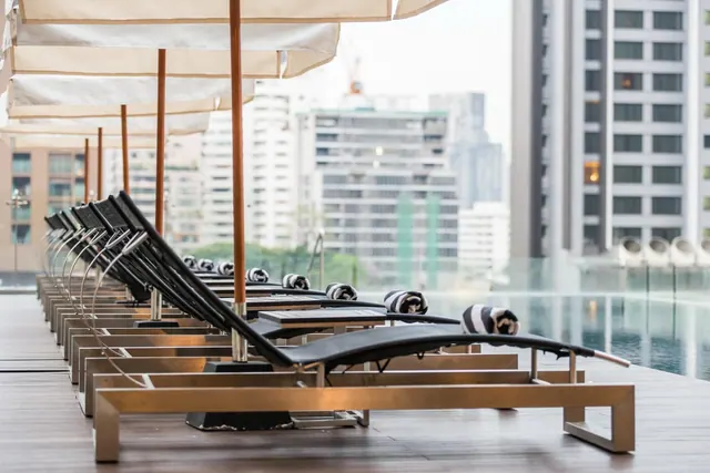 Rows of black sun loungers with striped towels and cream umbrellas sit on a wooden deck beside an infinity pool, with high-rise buildings in the background.