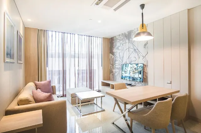 A living and dining area with a sofa and cushions, a wall-mounted television, a wooden dining table with four chairs, and floor-to-ceiling sheer curtains by the window.