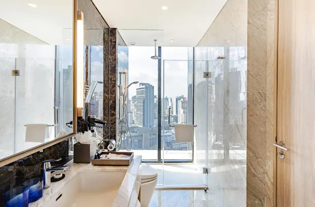 A marble bathroom with a glass-walled walk-in shower, an undermount sink with blue glasses, and a window behind the shower looking out over city high-rises.