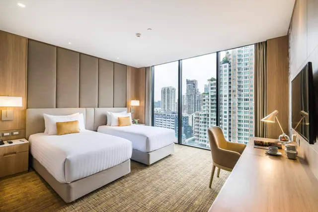 A hotel bedroom with two single beds, a wooden work desk and a wall-mounted television, with floor-to-ceiling windows looking out over high-rise residential towers.