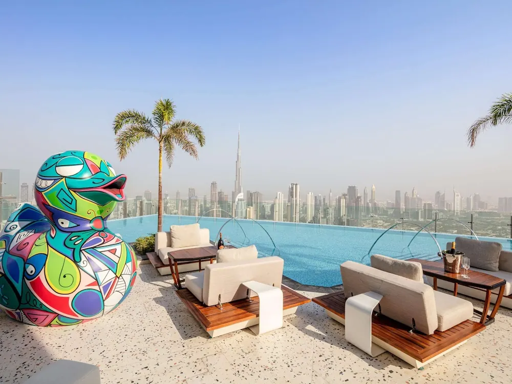 A rooftop infinity pool with lounge seating, a large colourful duck sculpture and a city skyline with the Burj Khalifa in the background.