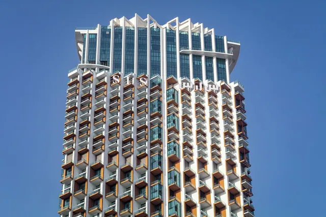 A high-rise tower with protruding balconies and the letters SLS on its upper facade, photographed against a clear blue sky.