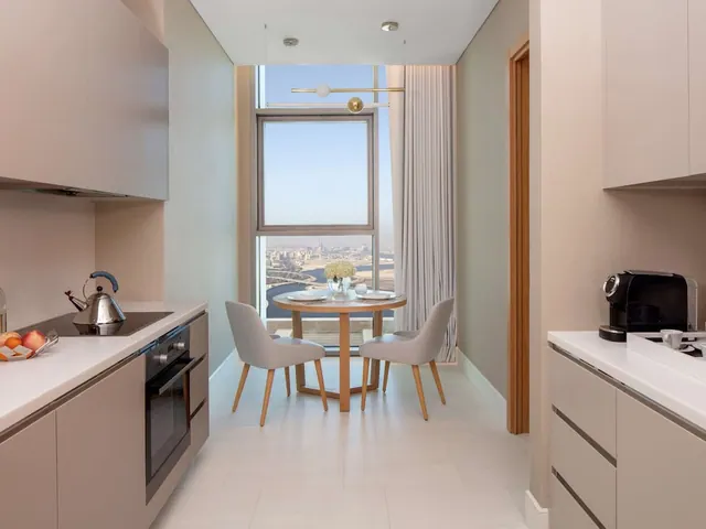 A galley kitchenette with a round dining table and two chairs sits in front of a tall window that looks out over water and a city skyline.