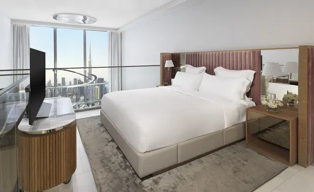 A bedroom with a king bed, floor-to-ceiling windows looking out over the city skyline including the Burj Khalifa, and a television on a wooden credenza.