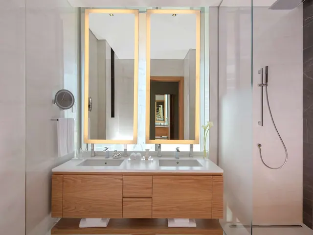 A bathroom with a wood double vanity under two backlit mirrors, a glass-walled walk-in shower and folded white towels.
