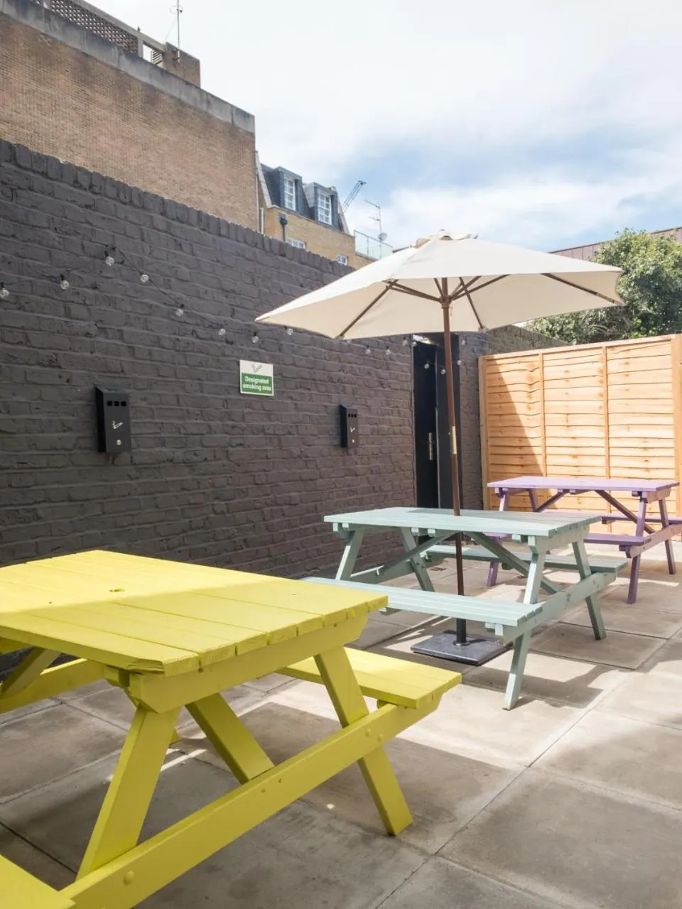 A paved outdoor terrace with yellow, green and purple picnic tables, a cream parasol, and a dark brick wall with a designated smoking area sign.