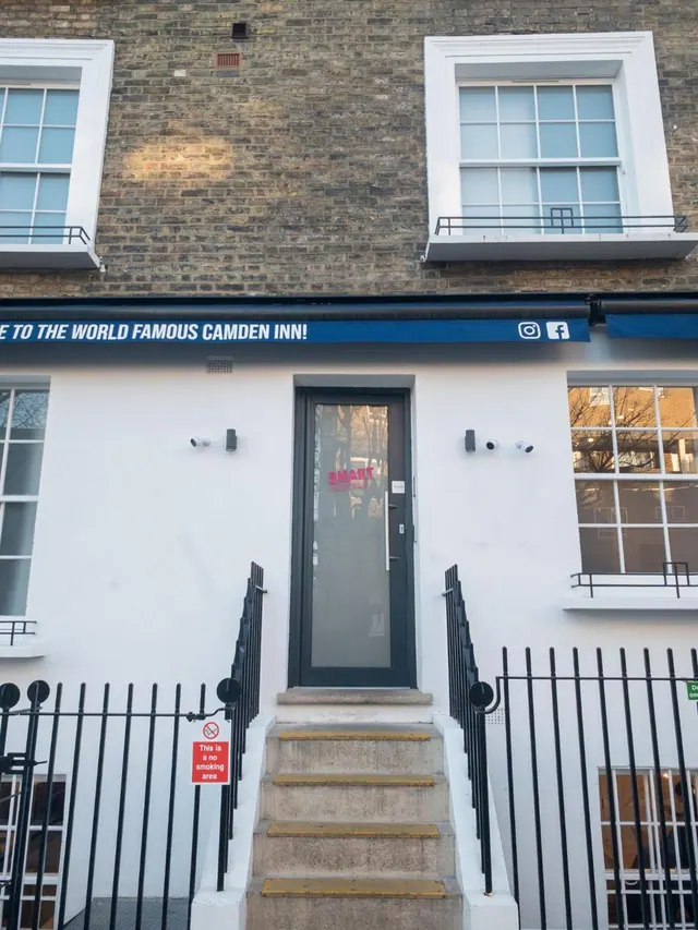 Street-level entrance of a white-painted brick building with a dark door, stone steps, black railings and a blue awning sign reading 'to the world famous Camden Inn'.