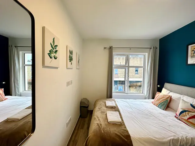 A bedroom with a double bed, folded towels on a brown throw, a teal accent wall, a wall mirror, and a window looking onto a brick building opposite.