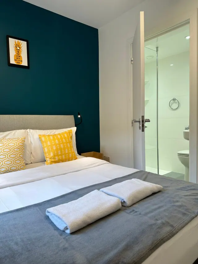 A double bed with a teal accent wall, yellow cushions and folded towels, with an open door to a bathroom containing a walk-in shower and toilet.