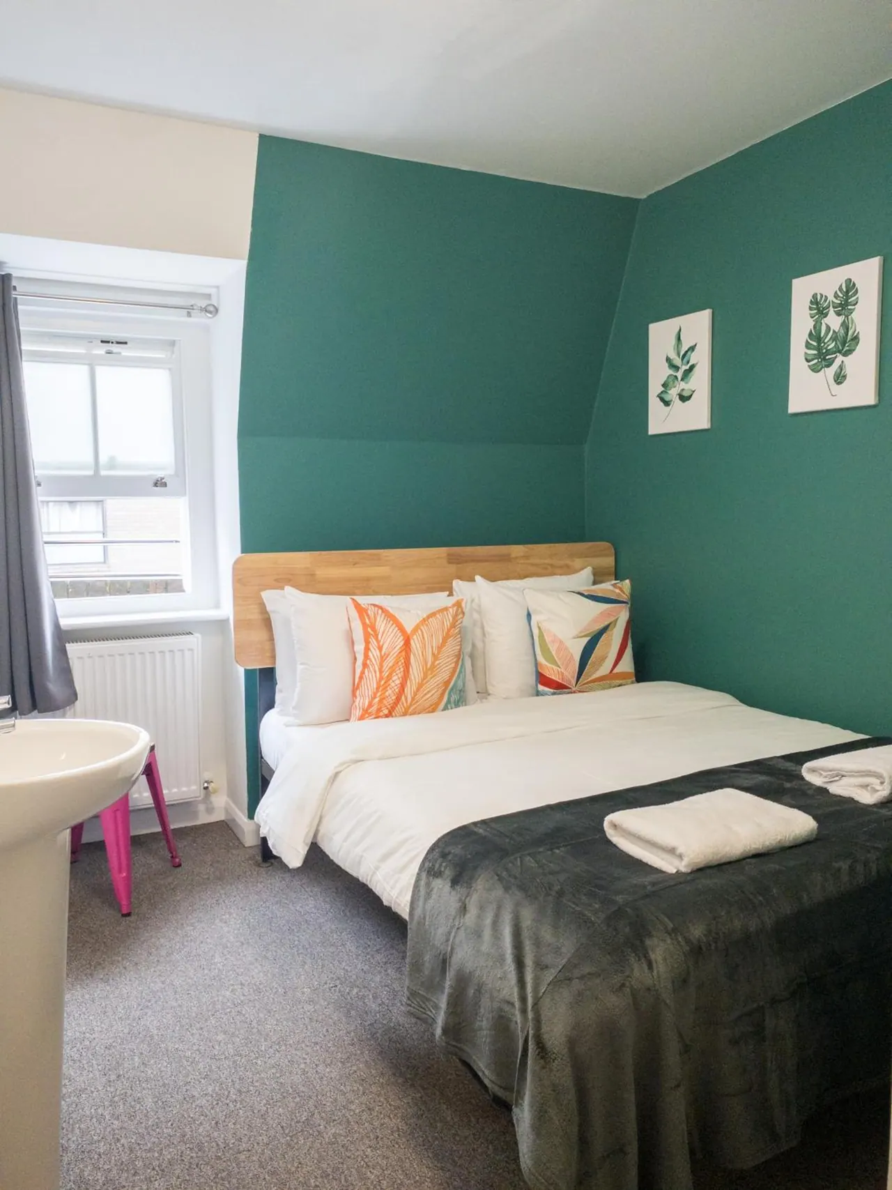 A bedroom with teal walls, a double bed with a wooden headboard and grey throw, a window looking onto a nearby building, and a pedestal sink at the left edge.