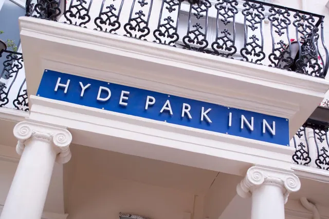 Looking up at the front of the Hyde Park Inn, showing a blue sign, white Ionic columns and black wrought iron balcony railings.