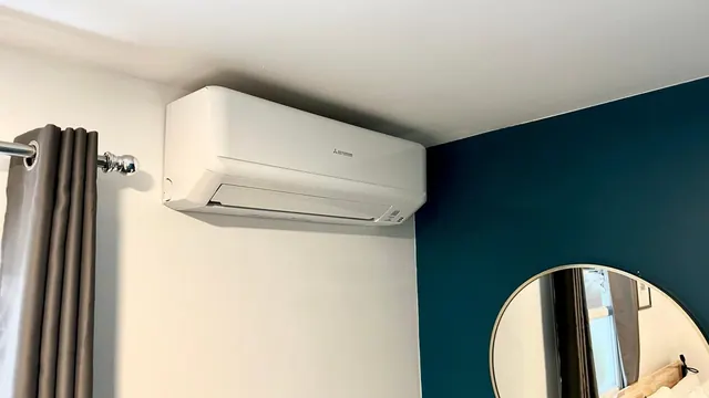 A white wall-mounted air conditioning unit is fixed to a pale wall above a grey curtain, beside a teal accent wall and a round mirror in a bedroom.