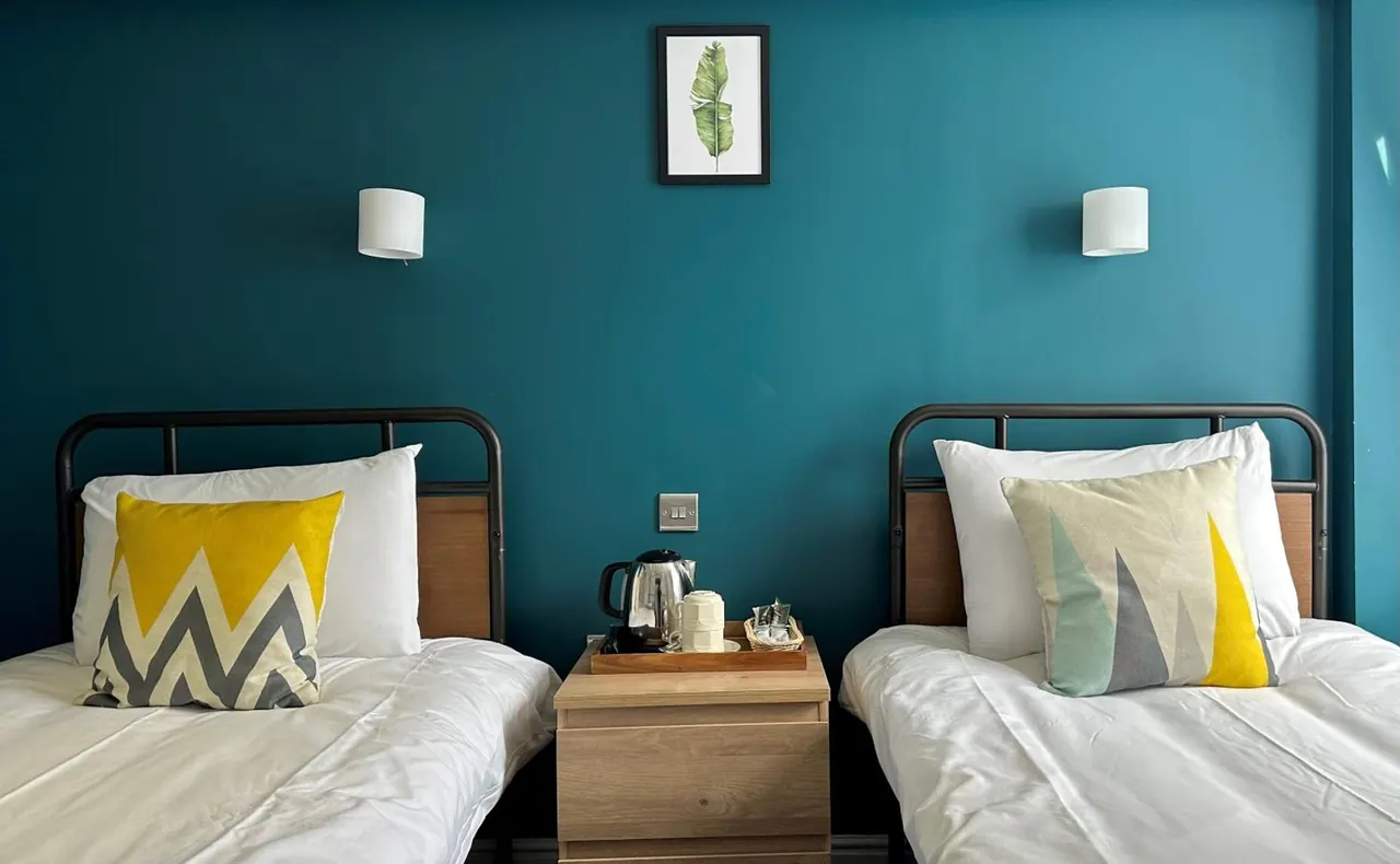 Two single beds with white linen and patterned cushions stand against a teal wall, with a bedside table holding a kettle and cups.