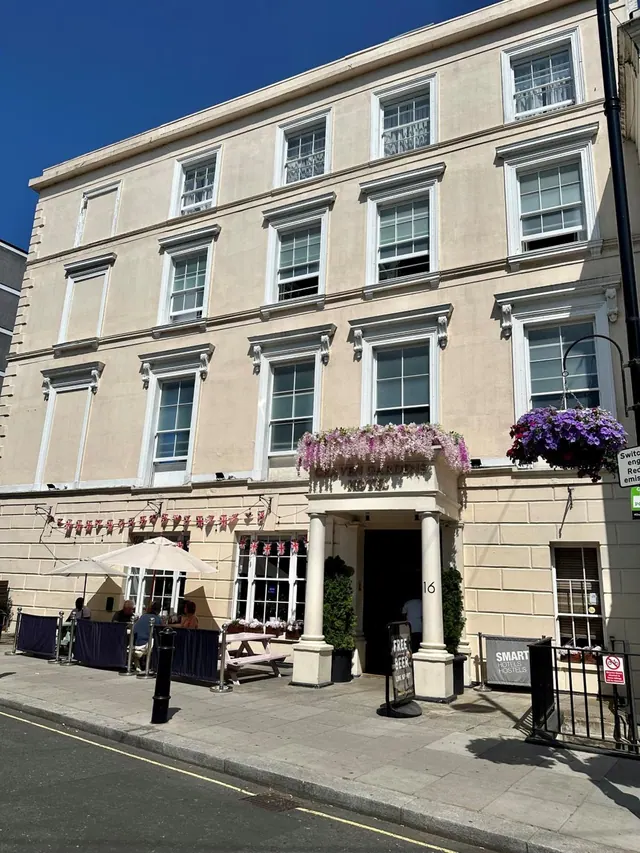 A cream-coloured multi-storey building with a columned entrance porch, hanging flower baskets and outdoor tables with umbrellas on the pavement beside it.