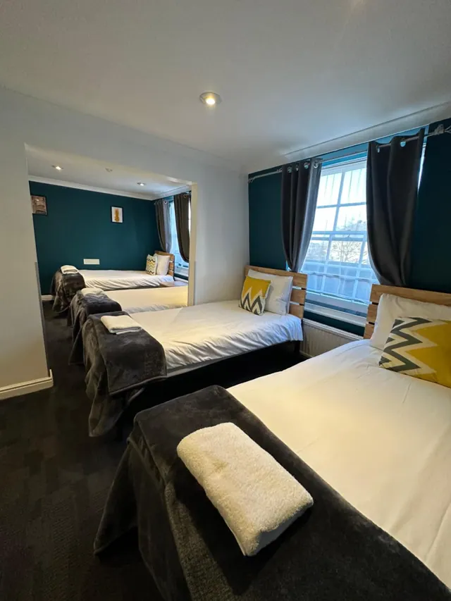 A bedroom with several made-up beds with white bedding, grey throws and yellow patterned cushions, beside a window with grey curtains and teal walls.