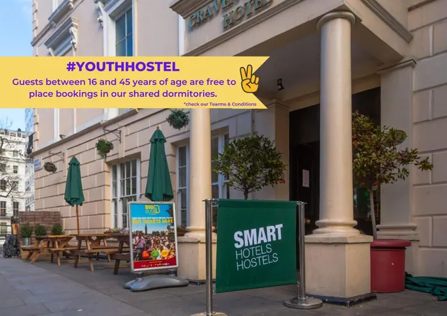 Front of a cream-coloured building with a columned entrance, green-umbrella picnic tables on the left, and a Smart Hotels Hostels sign on the pavement, with a yellow promotional banner across the top left.