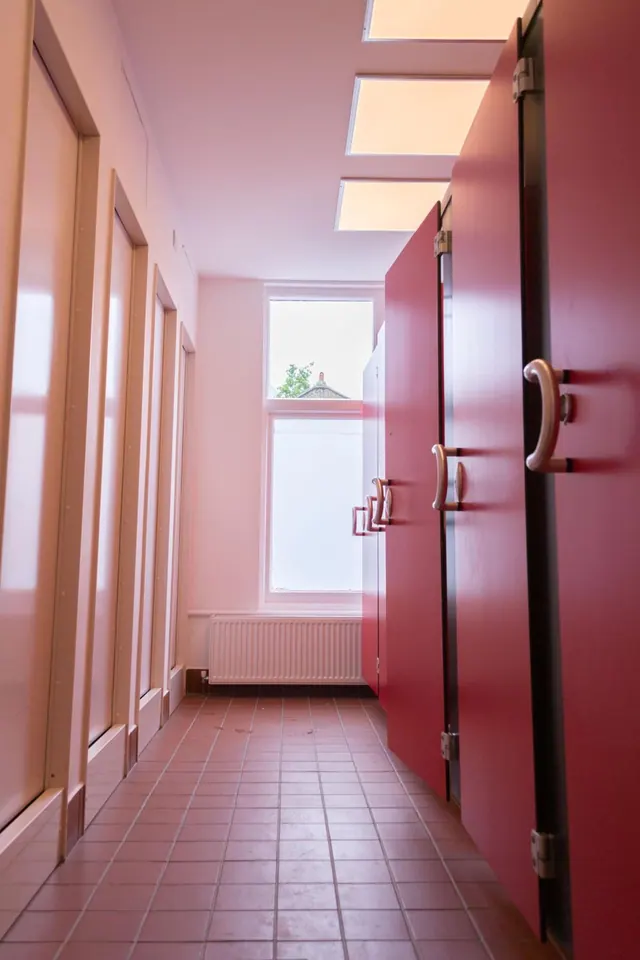 A tiled corridor lined with red and cream cubicle doors runs toward a window with a radiator beneath it, with skylights overhead.