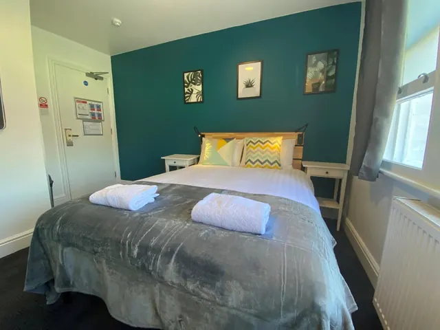 A bedroom with a double bed under a grey throw, two folded white towels on the bed, a teal accent wall, and a door with notices on the left.